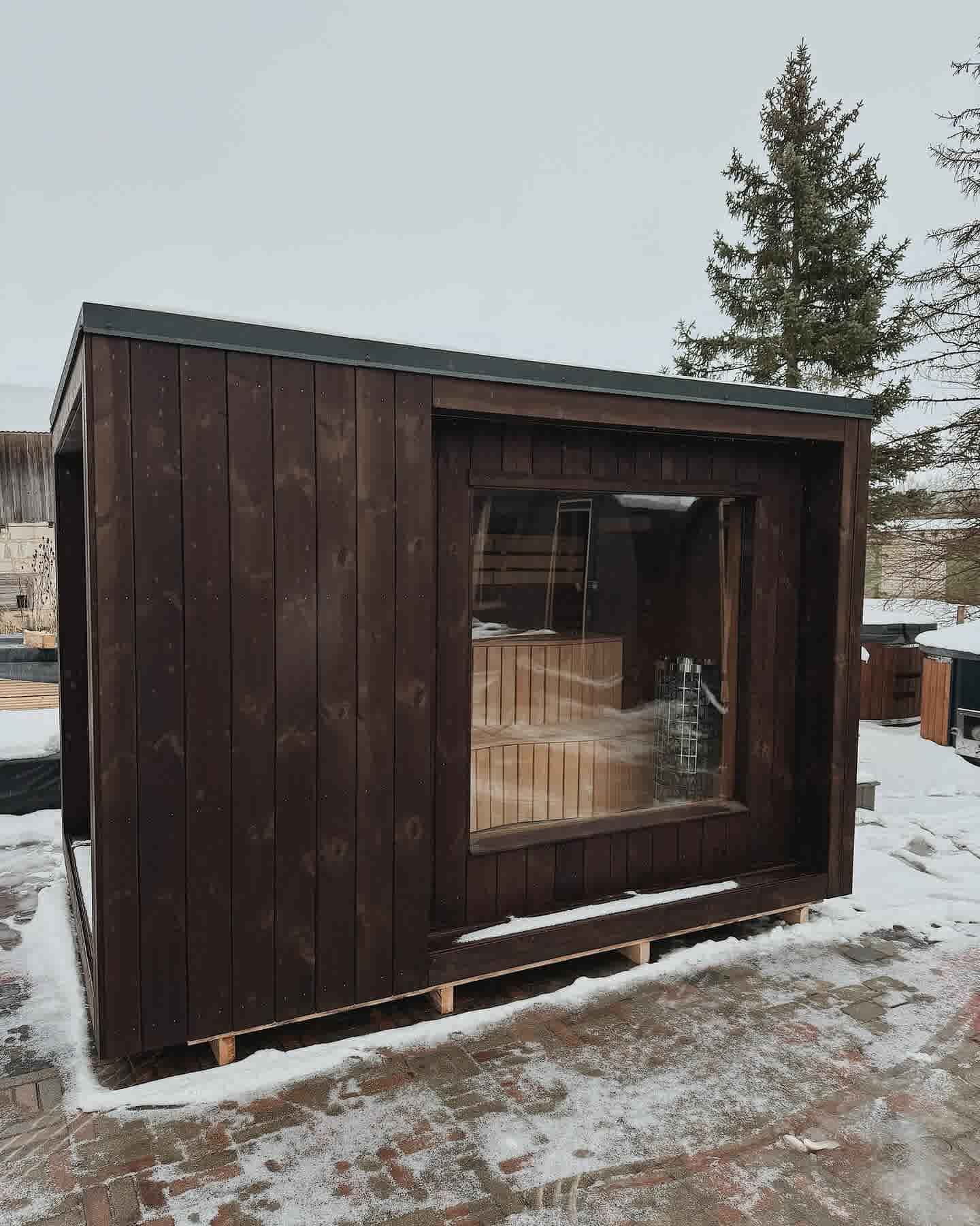 Exterior of a dark-stained modern cube sauna with a large panoramic window in a winter setting.