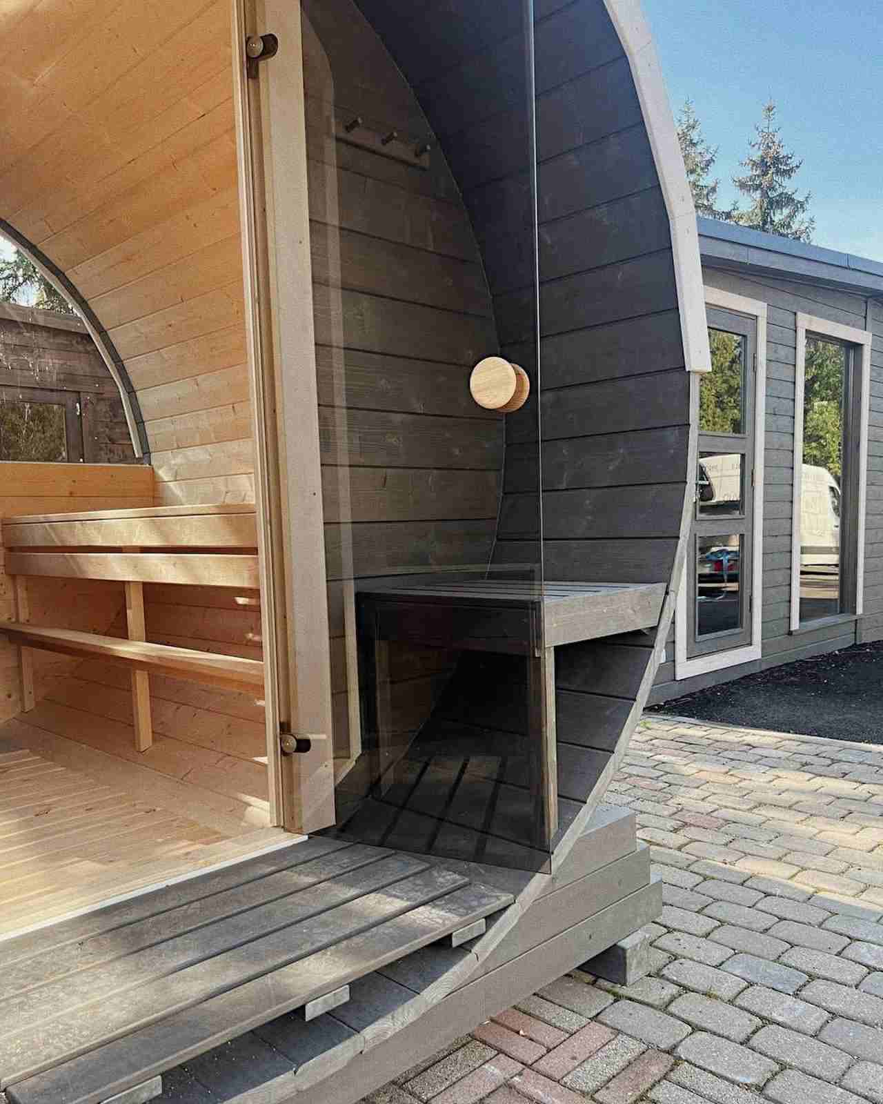 Side profile of a grey barrel sauna showing the glass door and external seating bench.