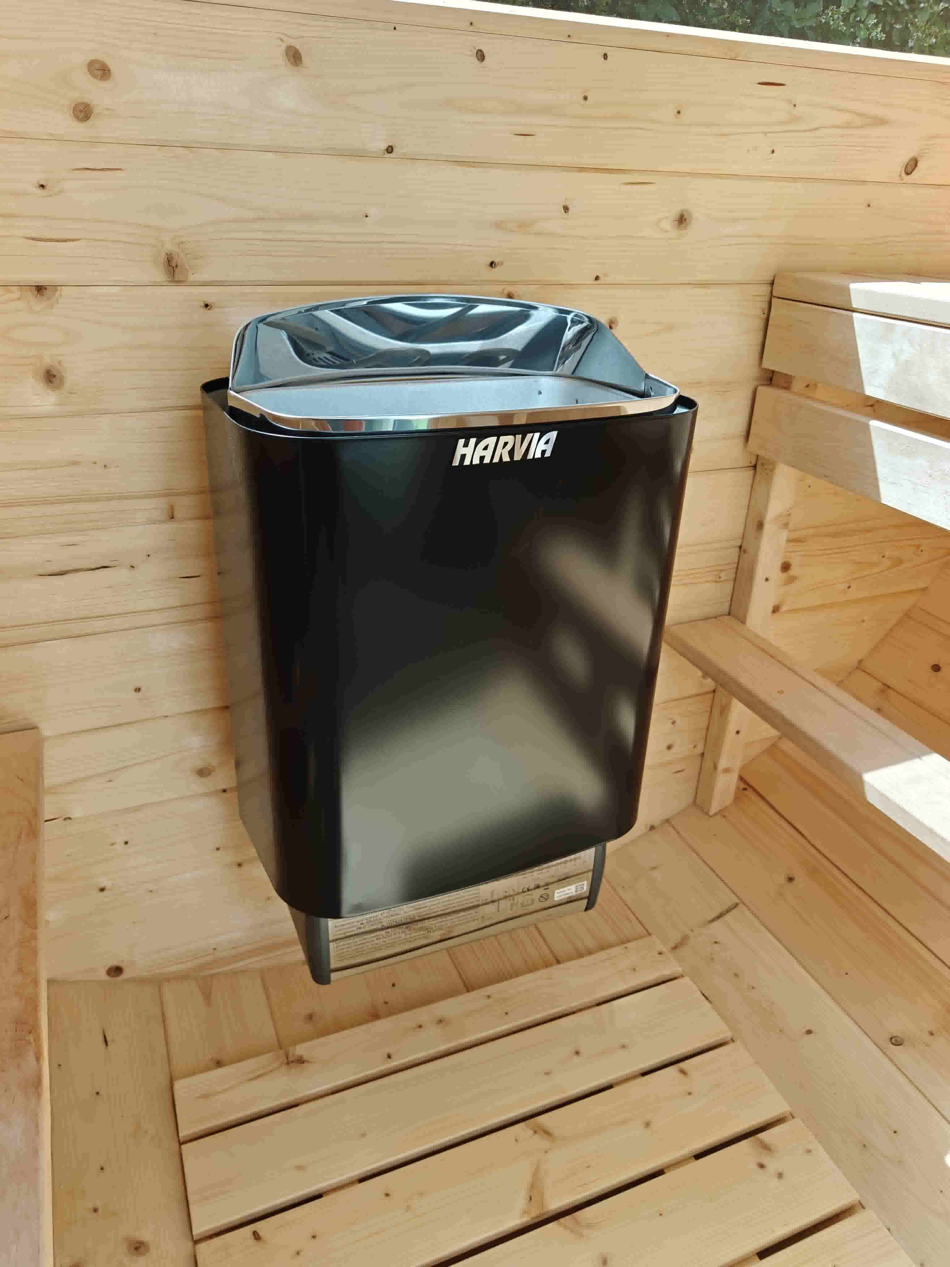 A sleek black wall-mounted Harvia electric sauna heater with a chrome top casing, set against natural wood-paneled walls and a slatted timber floor inside a Terra Sauna unit in Guernsey.