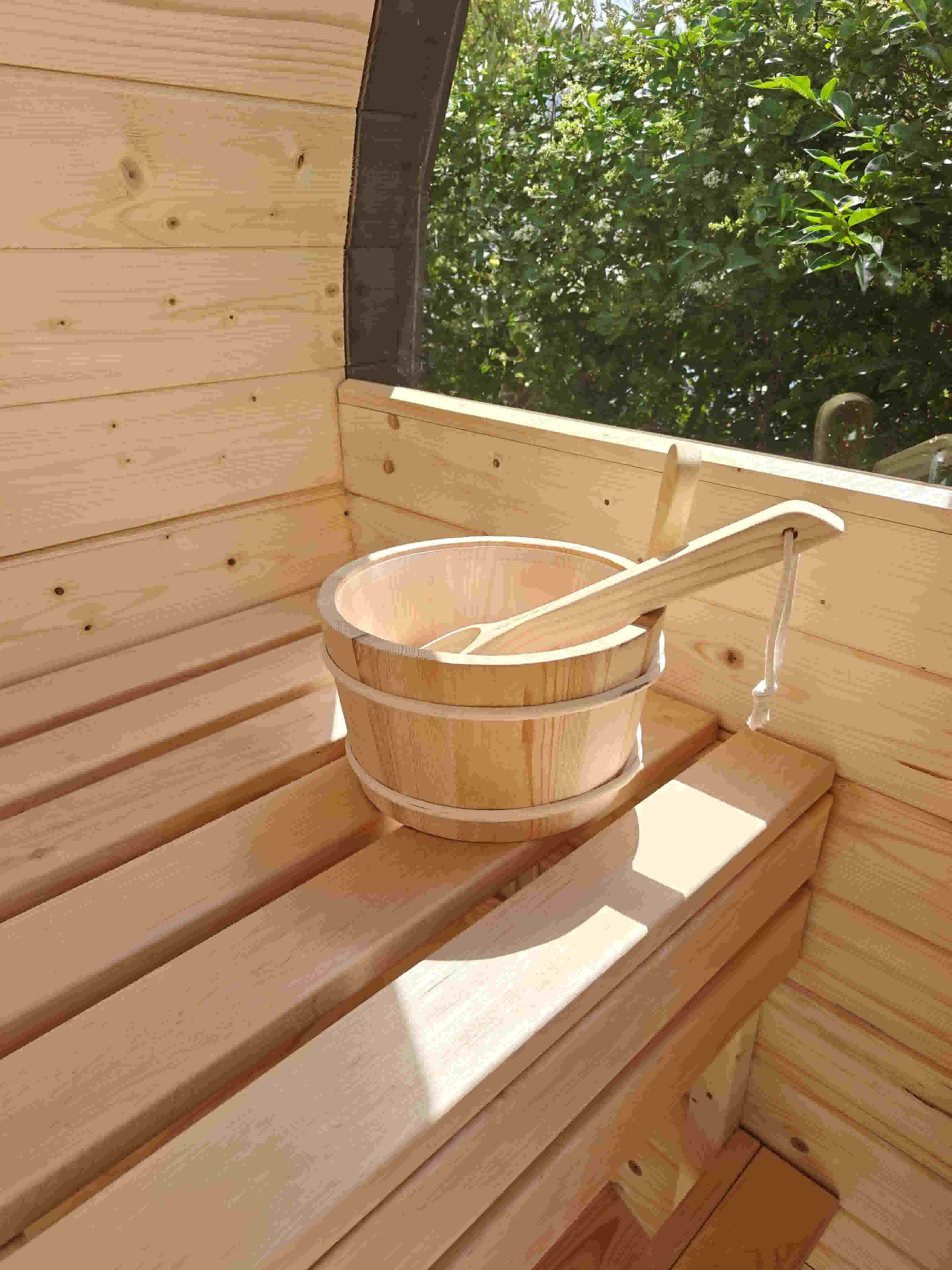 A classic wooden water bucket with a ladle resting on a sunlit alder bench next to a large panoramic window overlooking green hedges in Guernsey.